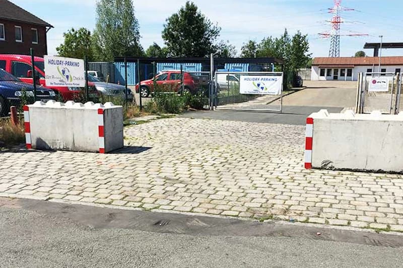 Einfahrt zu umzäuntem Parkplatz mit Betonbarrieren, Kopfsteinpflaster und offener Metallschranke.
