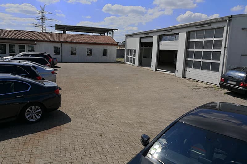 Gepflasterter Parkplatz mit mehreren geparkten Autos, Garagentoren rechts und Bürogebäude mit Ziegeldach im Hintergrund.