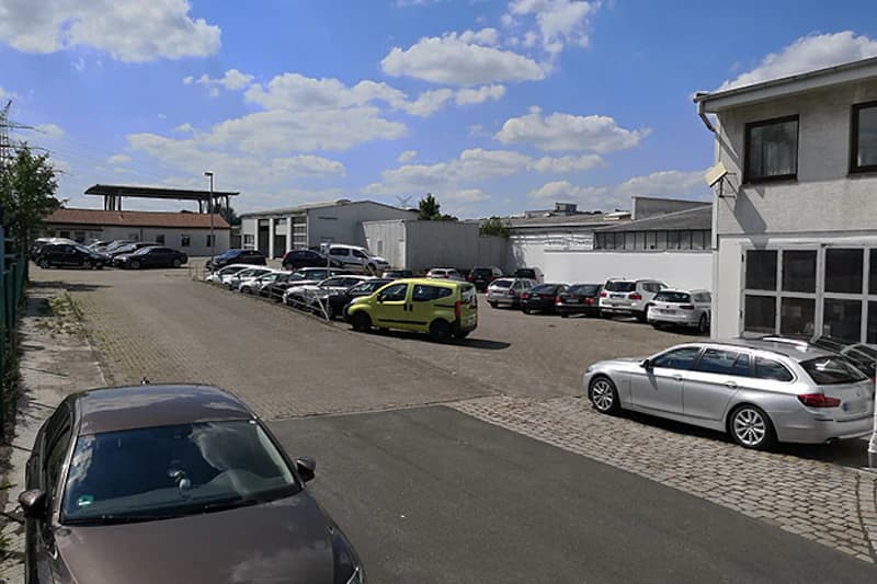 Offener Parkplatz mit geparkten Autos und niedrigen Hallen unter blauem Himmel mit Wolken.
