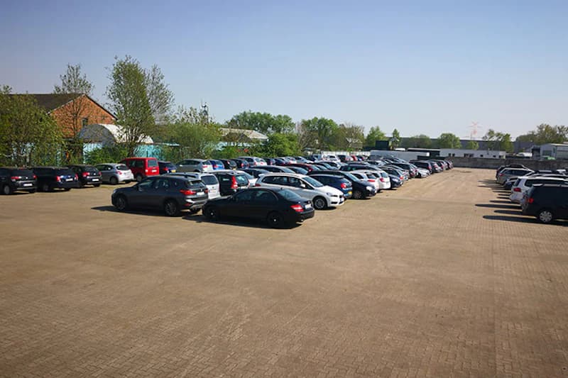 Gepflasterter Parkplatz am Flughafen Bremen mit mehreren dicht geparkten Autos, Bäumen und Gebäuden im Hintergrund.