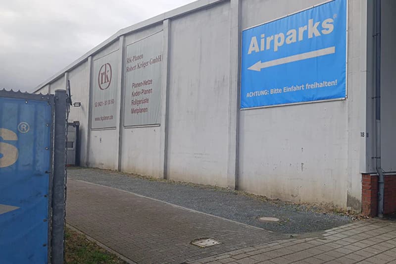 Außenwand eines Industriegebäudes mit blauem Airparks-Banner, Kiesfläche und gepflastertem Gehweg.