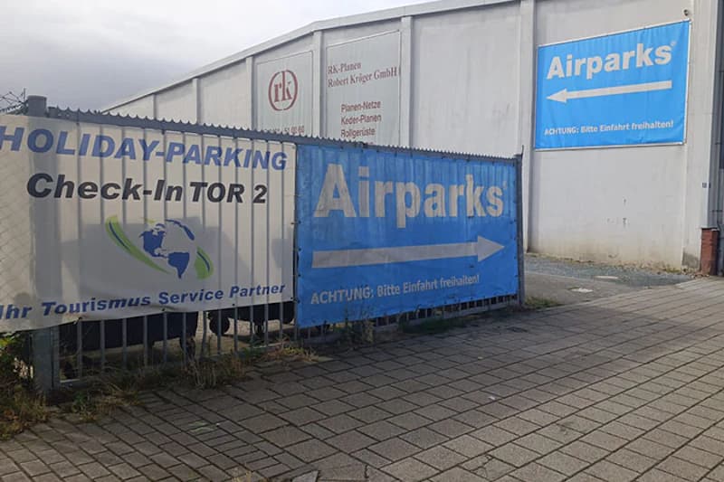 Banner an einem Zaun: Holiday-Parking Check-In TOR 2 und Airparks mit Richtungspfeil vor einer Halle.