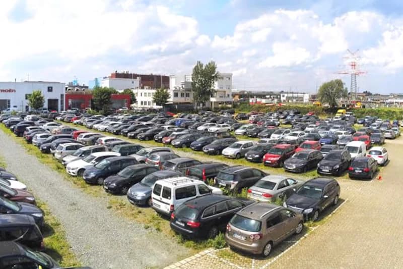 Großer eingezäunter Außenparkplatz mit zahlreichen geparkten Autos in Reihen, Kies- und Pflasterflächen, Flughafenbauten im Hintergrund.