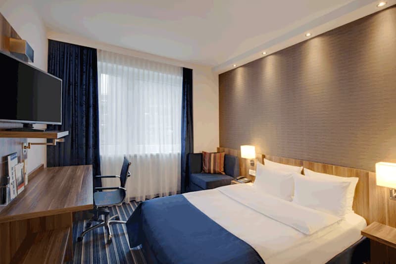 Hotelzimmer mit Doppelbett, zwei Nachttischen, Wandlampen, Fernseher, Schreibtisch, Bürostuhl und Fenster mit Vorhängen.