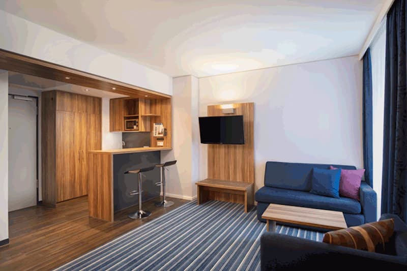 Hotelzimmer mit Sitzecke, blauem Sofa, Couchtisch, Wandfernseher und Holzkochnische mit Bartresen.