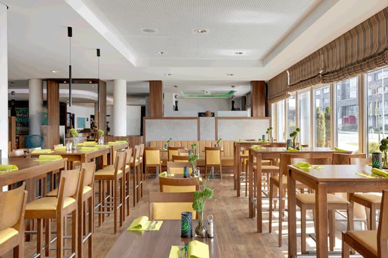 Heller Frühstücksraum mit Holztischen und hohen Stühlen, grüne Servietten und große Fenster rechts.