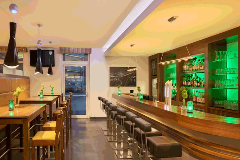 Hotelbar mit langem Holztresen, Barhockern, hohen Tischen, Hängeleuchten und grün beleuchteten Regalen.