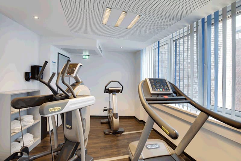 Kleiner Fitnessraum mit Laufband, Crosstrainer, Fahrradtrainer, Regal mit Handtüchern und großen Fenstern.