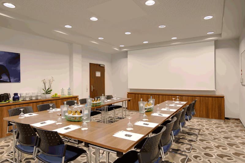 Konferenzraum mit U-förmigen Tischen, Stühlen, Schreibunterlagen, Wasserkaraffen und Leinwand.